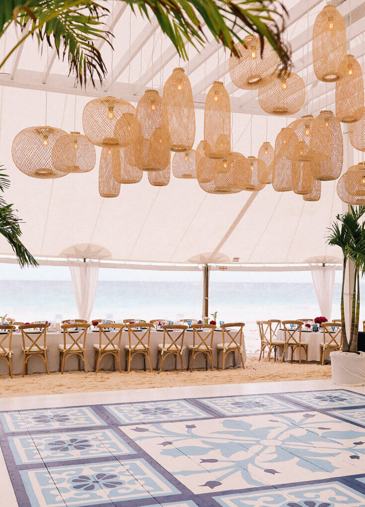 Ceiling Wedding Decor: Mismatched rattan light fixtures hanging from the ceiling of a white oceanfront tent with a blue dance floor below.