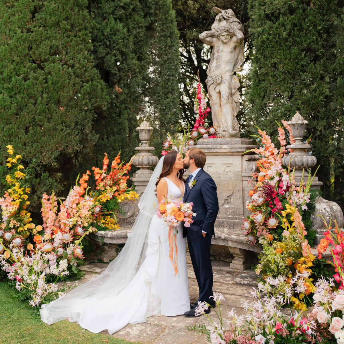 A bride and groom kissing in a garden decorated with colorful flowers around a statue at their Tuscan wedding ceremony.