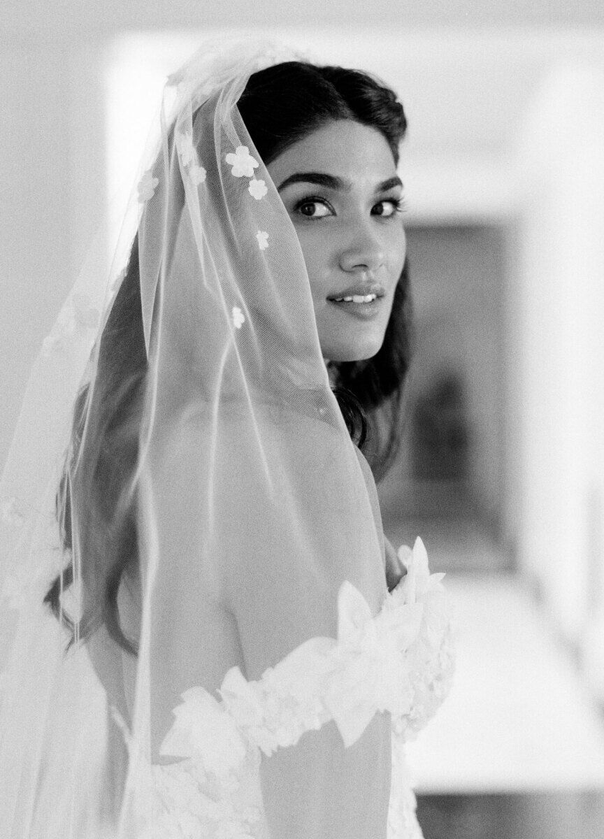 A bride, in a veil and off-the-shoulder wedding dress, looks at her photographer during a pre-ceremony portrait in her parents' home before her waterfront wedding began.