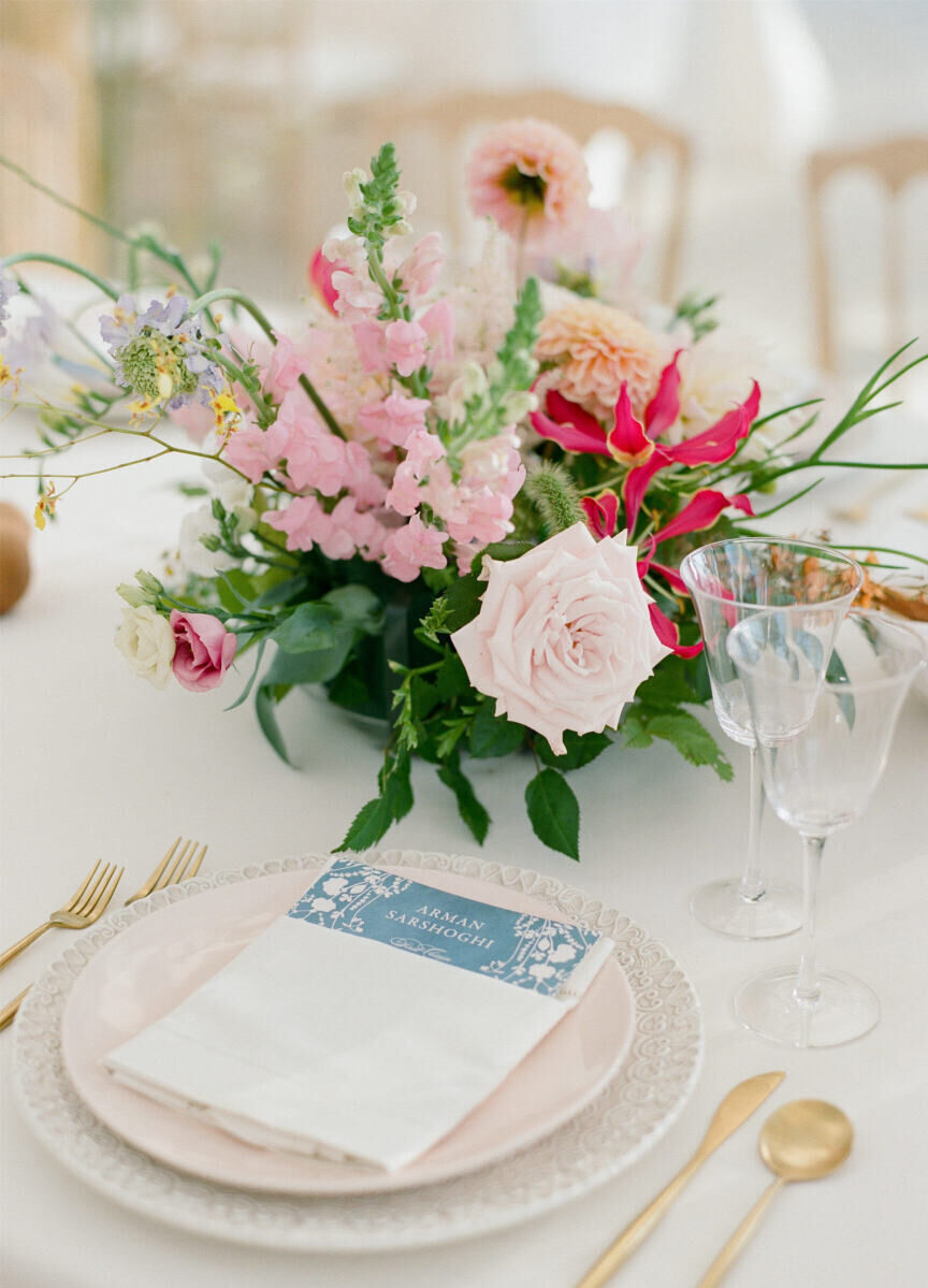 A centerpiece of assorted pink flowers decorates a table set with ornate white chargers, simple blush pink plates, modern gold flatware, and a floral-patterned dinner menu at a waterfront wedding in Long Island.