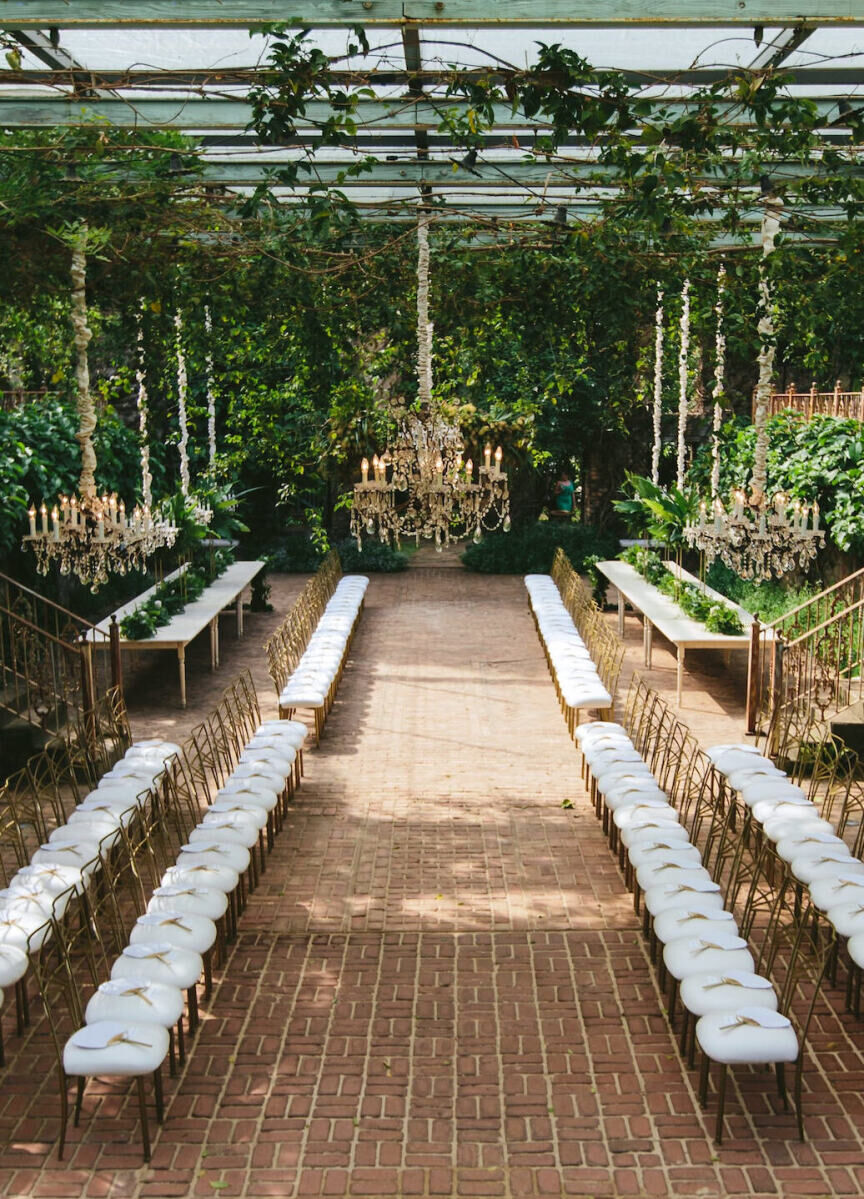 Wedding Aisle: An indoor-outdoor ceremony with two rows of runway-style cushioned chairs and a gold chandelier hanging above.