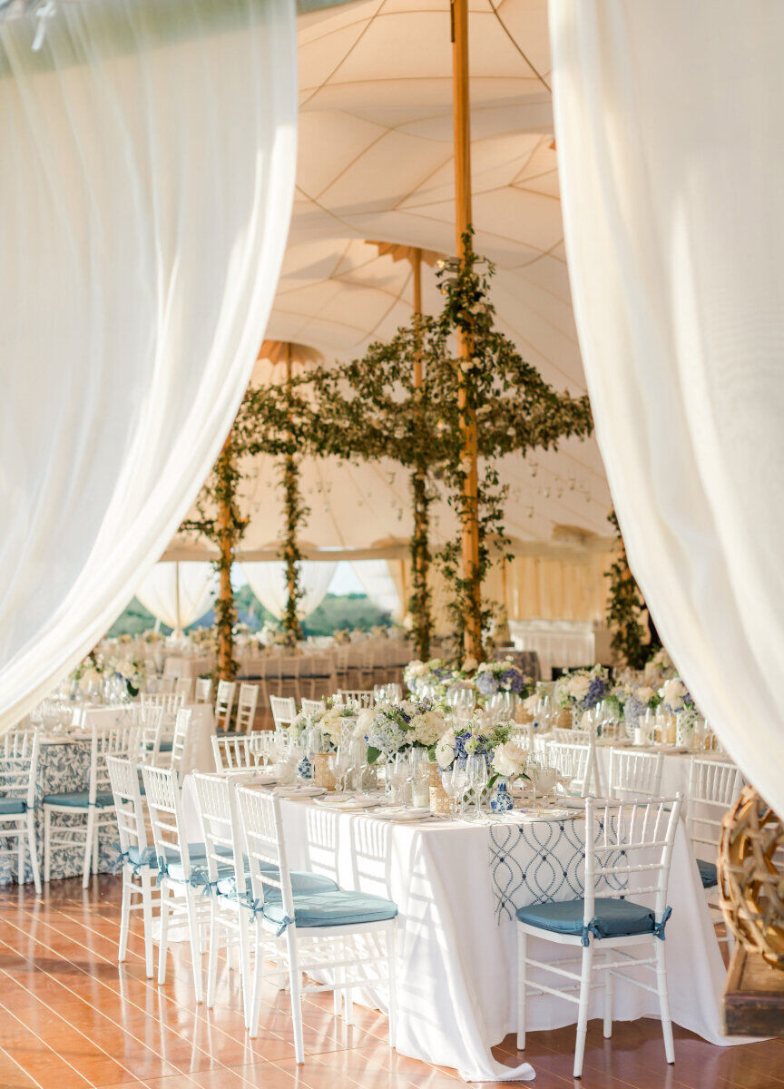 Wedding Tips: A white and blue reception set-up of tables and chairs in a big, airy white tent. The tent has green florals hanging from the poles.