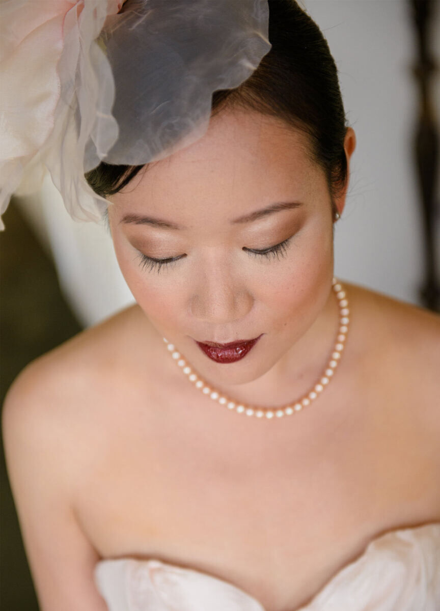 Wedding Tips: Close-up photo of a bride's makeup. The bride is looking down and is wearing a pearl necklace, with a dark maroon/red lip color.