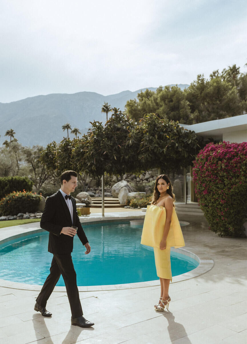 Wedding Tips: A bride in a yellow dress with a groom walking by in a tux. They are both standing by an outdoor in-ground pool and there are mountains and trees in the background.