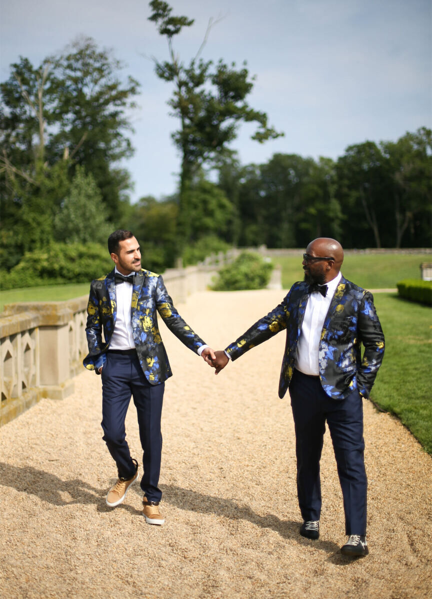 Two grooms in colorful, floral jackets and suits holding hands outside.