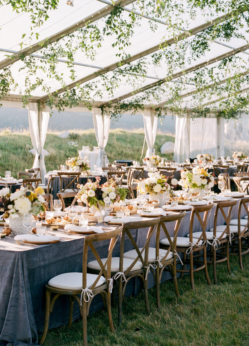 Wedding Tips: A reception set-up with a long table under a tent in a private residence. There are flowers lining the table and greenery hanging from the roof of the tent.