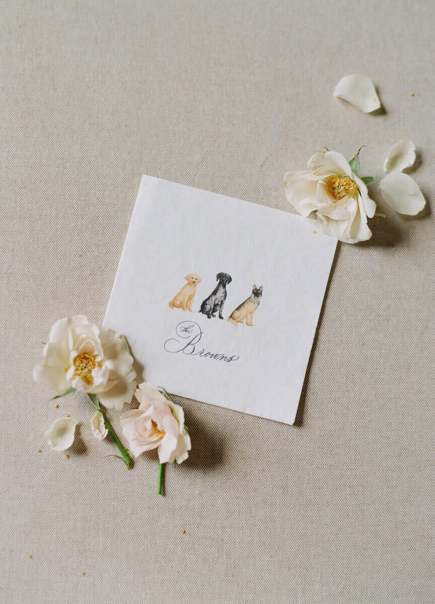 Wedding Tips: A cocktail napkin surrounded by floral blooms. The cocktail napkin has illustrations of three dogs with words that say, 