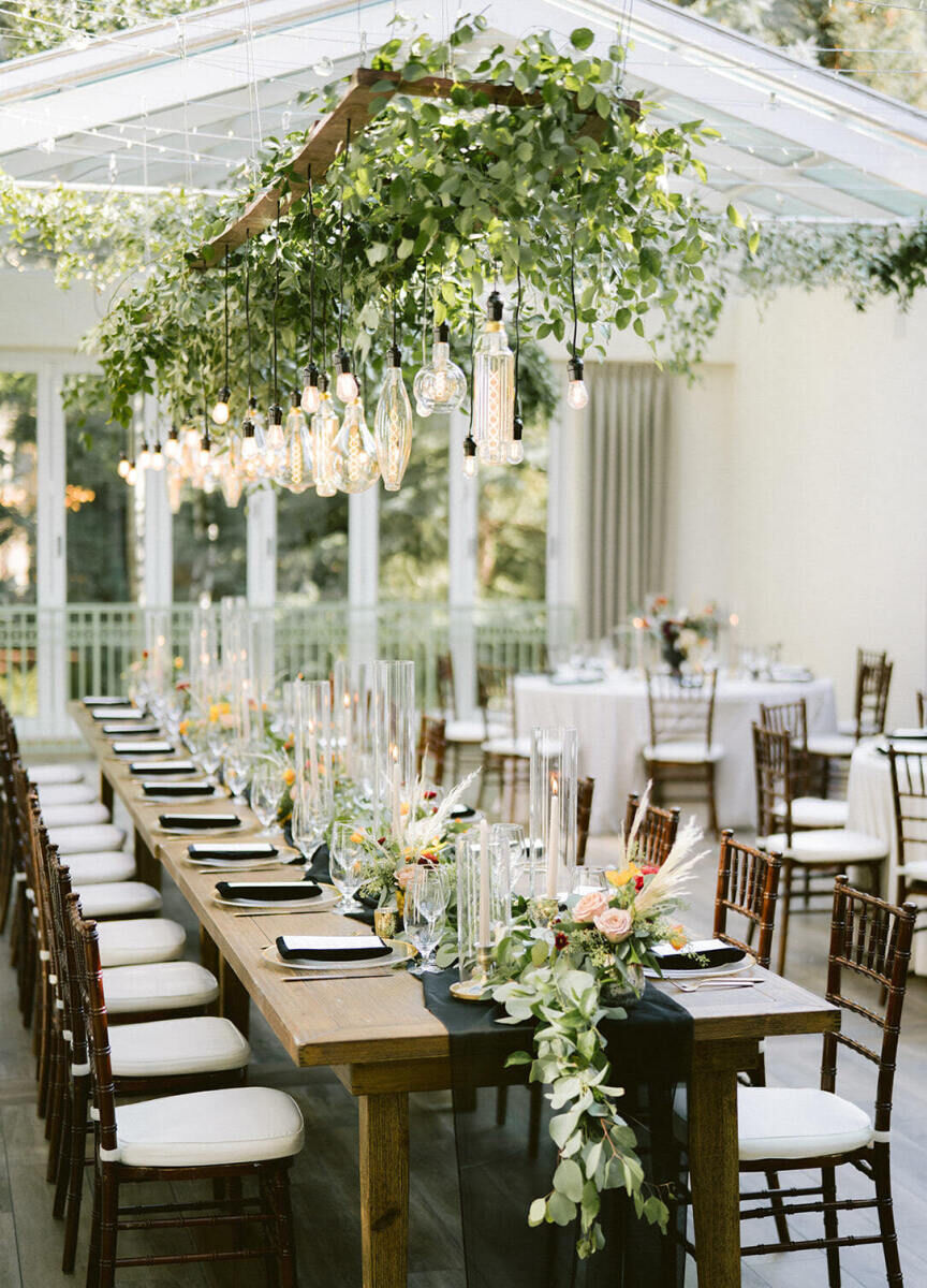 Wedding Tips: A reception set-up with a long table for guests with chairs on both sides and greenery hanging above.