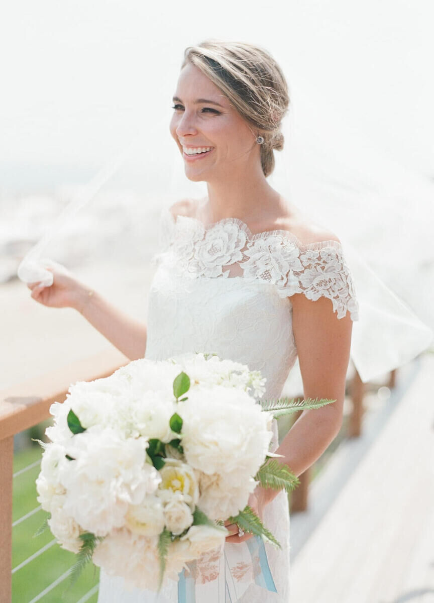 Wedding Tips: A bride in a white, strapless gown with a lace overlay smiling and holding her veil and a bouquet of white flowers.