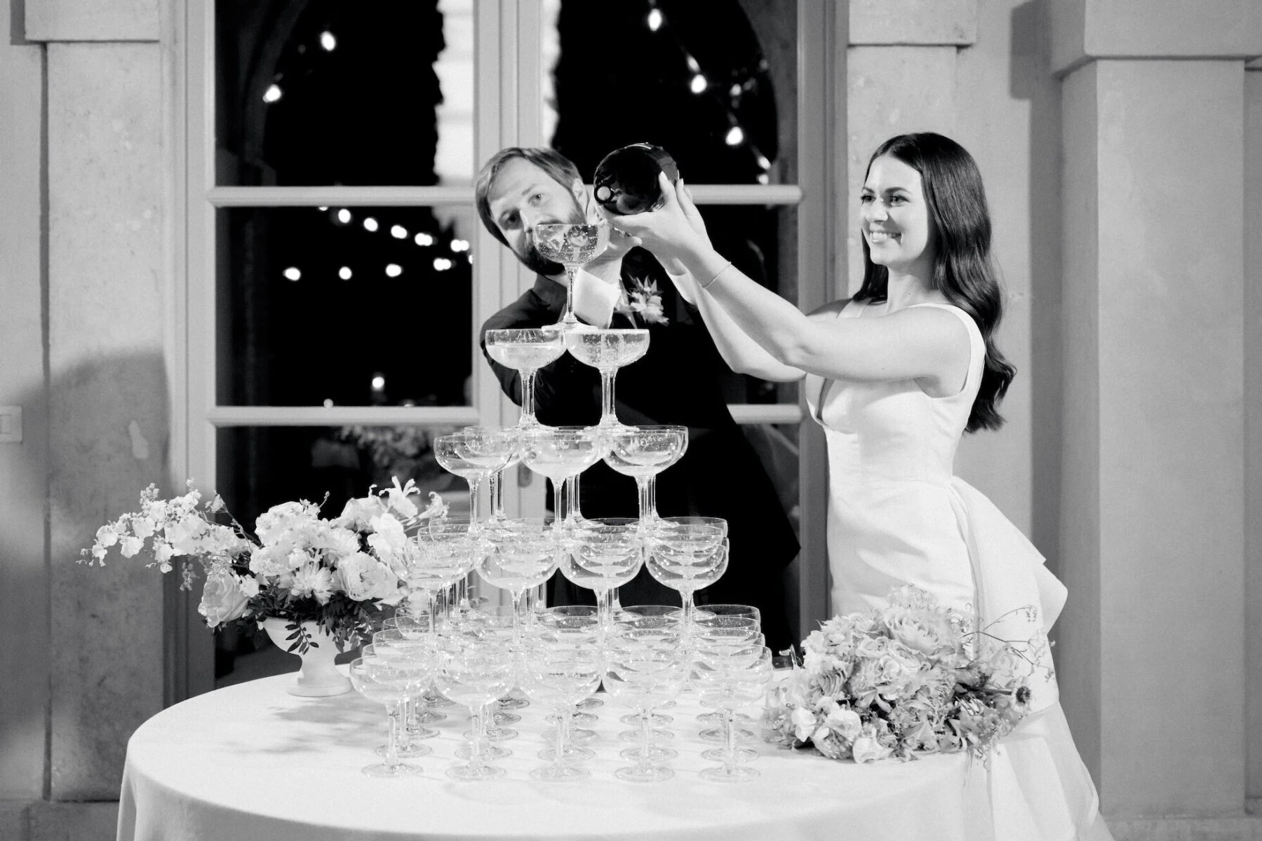 A groom and bride pour champagne over a tower of coupes at their Tuscan wedding reception.