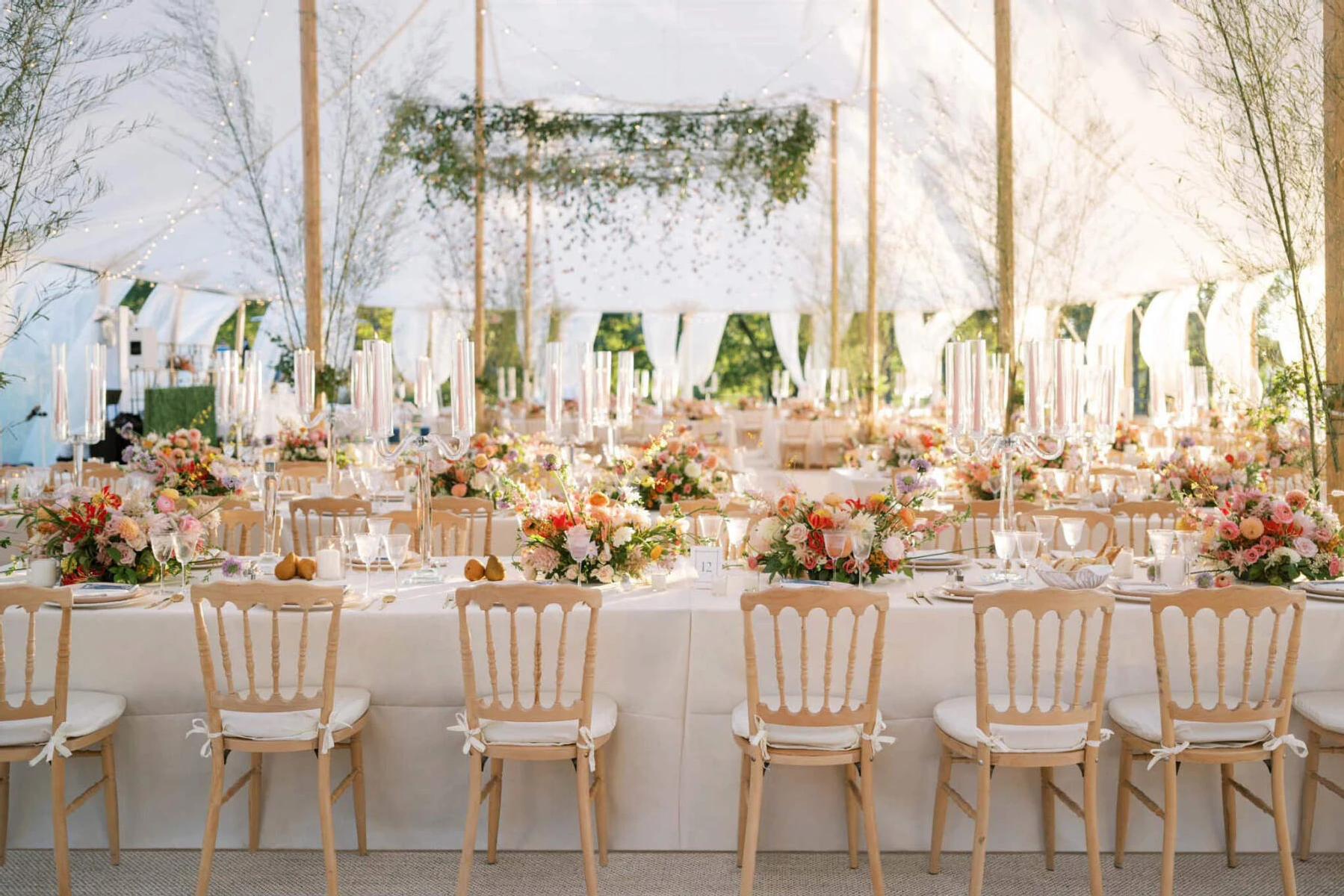 The tented reception of a waterfront wedding, decorated with a canopy of greenery and folded paper cranes over the dance floor and centerpieces made with a variety of warm-hued flowers and fruit, paired with elegant pink taper candles.